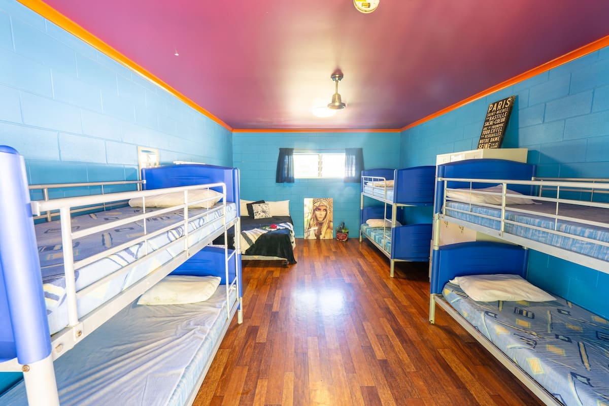 A dormitory room with bunk beds, wooden flooring, a ceiling fan, and a window with curtains.
