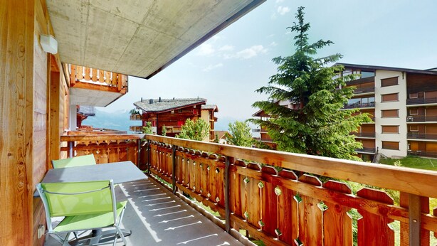 Ein Holzbalkon mit Tisch und Stühlen, der auf eine gebirgige Landschaft und ein Gebäude mit Balkonen blickt.