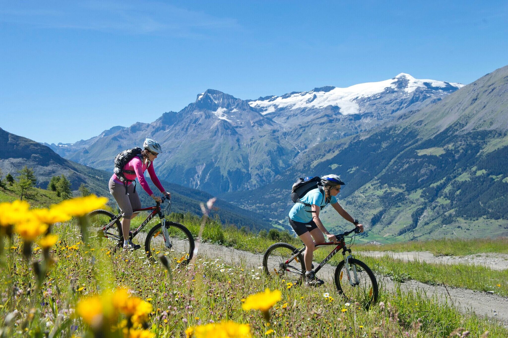 Two cyclists on a mountain trail with a snow-capped peak in the background.