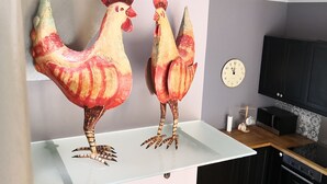 Twee grote, kleurrijke haanbeelden op een witte plank in een keuken met een magnetron, kastjes en een klok aan de muur.