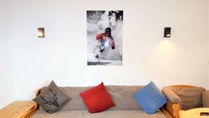 Een kamer met een bank, twee kussens, een houten nachtkastje en een aan de muur hangend schilderij van een skiër.