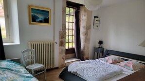 Ein Schlafzimmer mit Bett, Sessel, Heizkörper, Fenster mit Vorhängen, Lampe und einem Bild an der Wand.