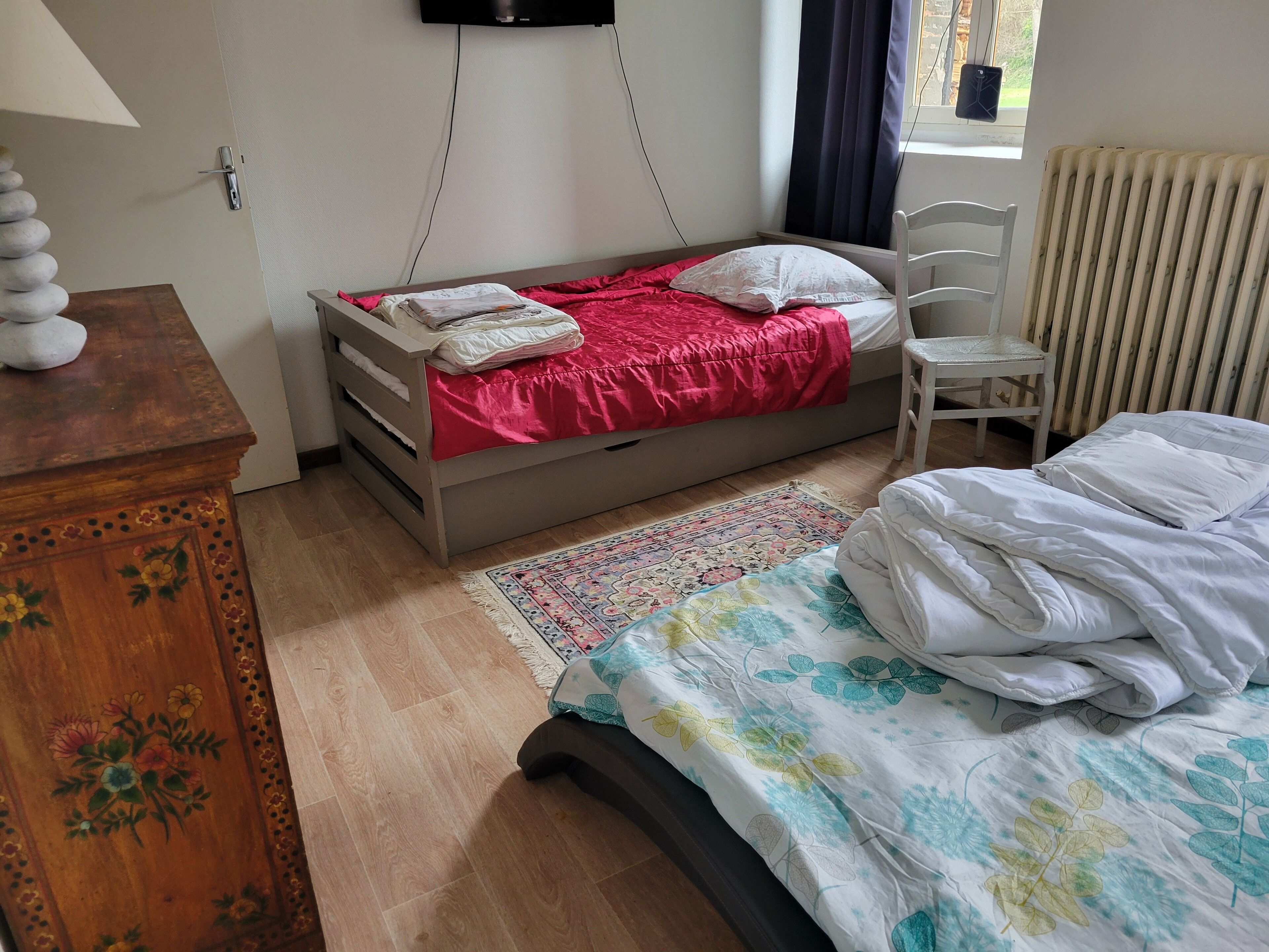 A bedroom with a bed, a chair, a dresser, a radiator, and a window with curtains.