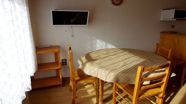 A dining area with a round table, wooden chairs, a television mounted on the wall, and a microwave on a cabinet.
