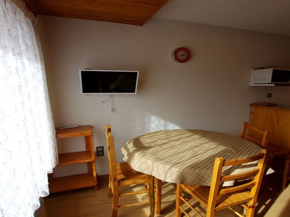 A dining area with a round table, wooden chairs, a television mounted on the wall, and a microwave on a cabinet.