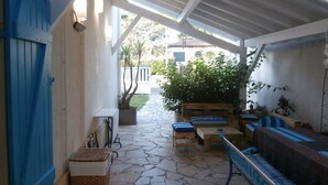 A covered outdoor seating area with stone flooring, wooden furniture, and a view of a white building in the background.
