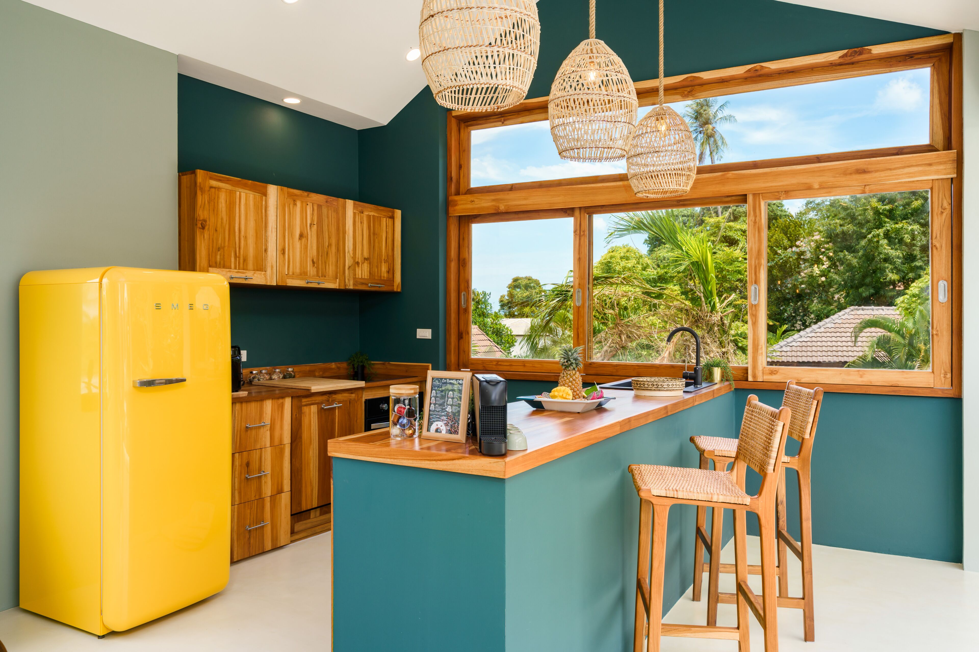 Una cocina con un refrigerador amarillo, armarios de madera, una isla de madera con taburetes de bar y ventanas amplias con vista a follaje.