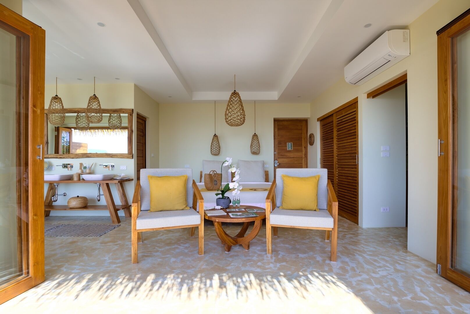 A spacious room with a wooden floor, two sofas with cushions, a small round table, a bed with a wooden headboard, a large mirror, and an air conditioning unit.