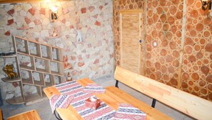 Una habitación rústica con pared de piedra, bancos de madera, mantel estampado y estante de madera con objetos decorativos.