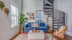 A living room with a blue sofa, a patterned rug, a wooden coffee table, a wicker chair, and a spiral staircase.