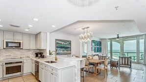 A modern kitchen with white cabinetry, a central island, and a dining area with a view of the ocean. - Tower 3- Amazing Gulf Views at Portofino 3-1909 (Pensacola Beach)