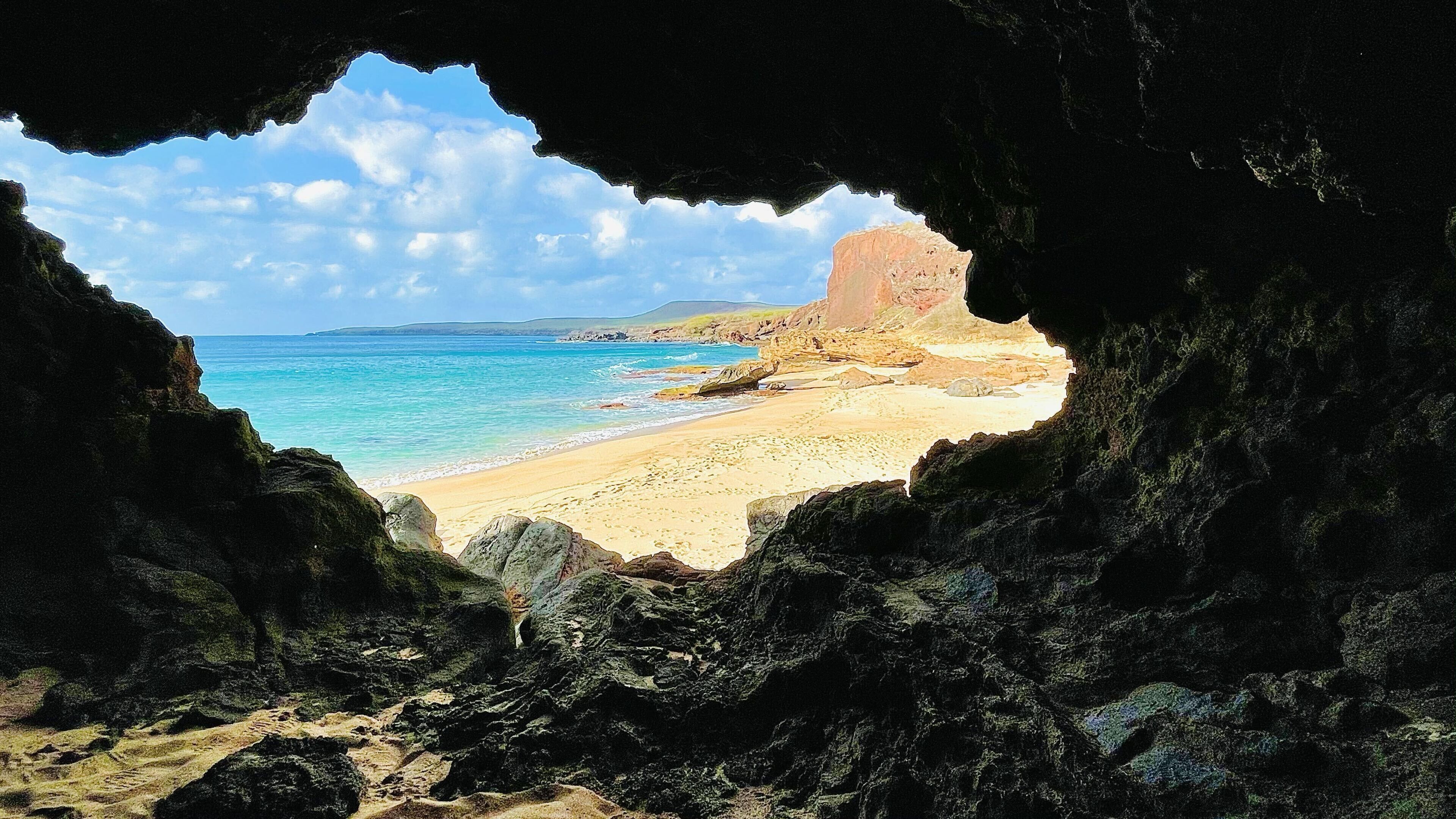 Une scène de plage, vue à travers l’entrée d’une grotte, avec une plage de sable, une eau bleue et limpide, et des formations rocheuses lointaines.