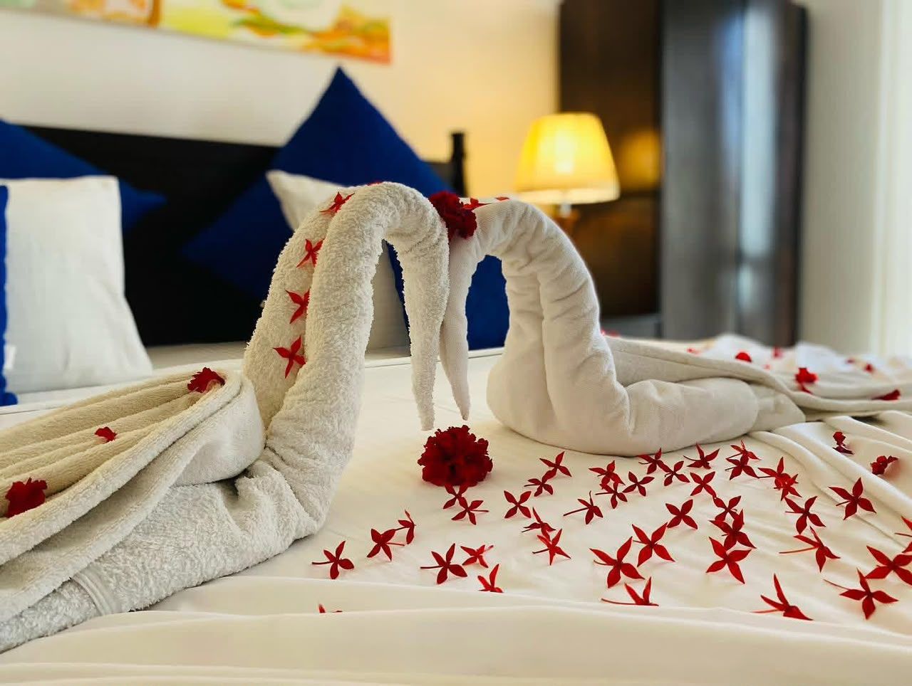 Habitación de hotel con una cama impecable, adornada con dos toallas con forma de cisne y pétalos de flores rojas.