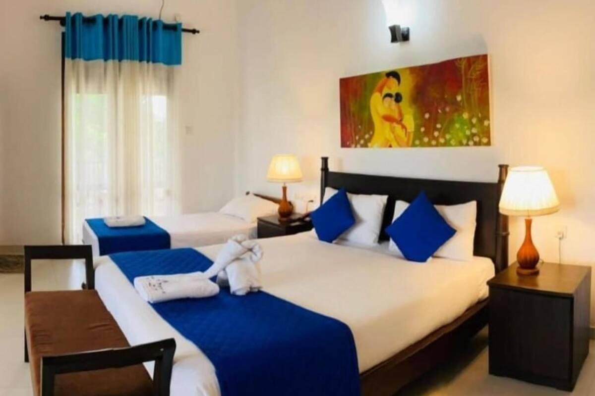 Een hotelkamer met twee bedden, elk met blauw beddengoed en witte kussens. Twee nachtkastjes met lampen, een schilderij boven de bedden, en een raam met gordijnen.