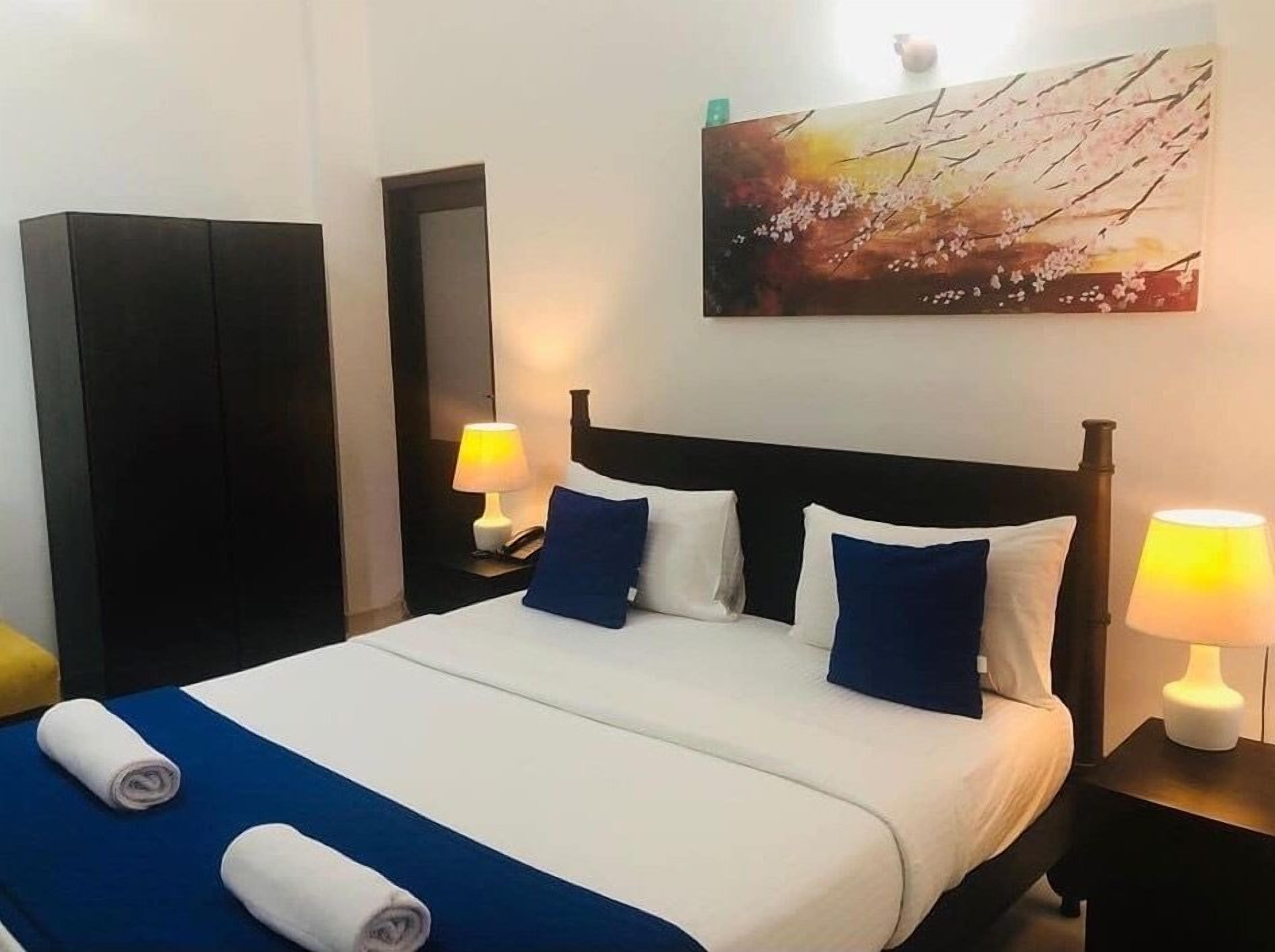 Ein ordentlich bezogenes Bett mit einer blau-weißen Tagesdecke, zwei Kissen und zwei gerollten Handtüchern. Darüber an der Wand hängt ein Bild, links befindet sich ein Kleiderschrank und rechts neben dem Bett jeweils eine Nachttischlampe.