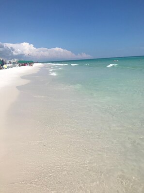 Une plage à l’eau turquoise et limpide, un rivage sablonneux et quelques personnes visibles au loin.