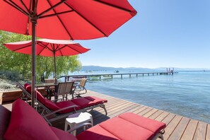 Eine Holzterrasse mit roten Liegestühlen und einem Tisch, mit Blick auf ein ruhiges Gewässer, das mit einem Steg endet.