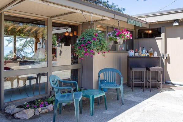 Eine Terrasse mit zwei blauen Plastikstühlen, einem hängenden Blumenkorb und einer Bar mit Flaschen.