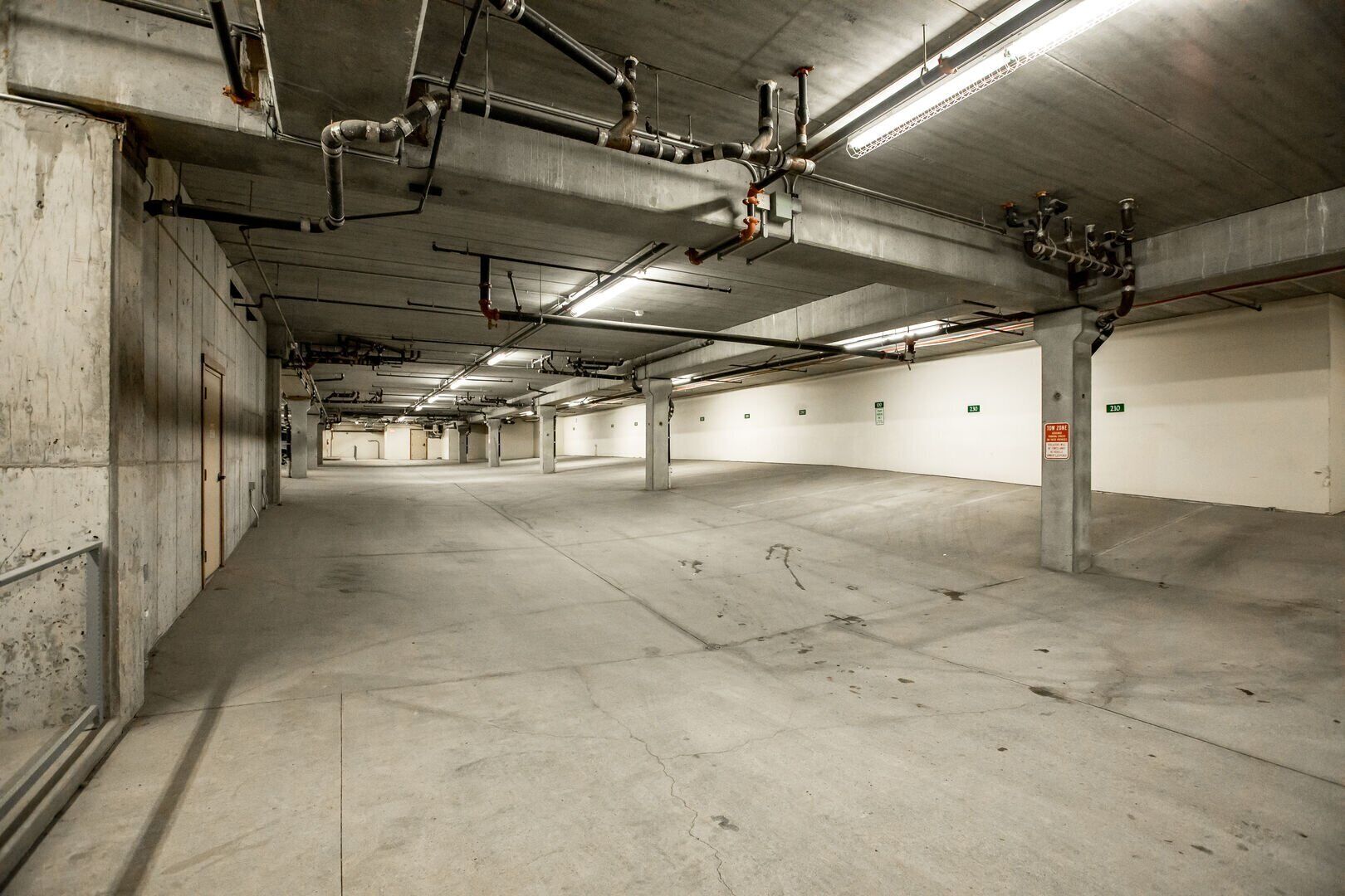 An empty parking garage with concrete floors and walls, visible structural supports, and overhead pipes.