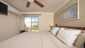 A hotel room with a large bed, a ceiling fan, a nightstand with a lamp, and a window with a view of buildings and greenery. - Oceanfront Condo on magic Sugar Beach - Ocean View (Kihei)