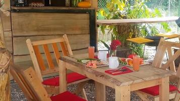 A wooden outdoor dining area with red cushions, a coffee maker, and a variety of fruits and flowers on the table.