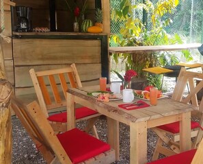 Uma área de refeições ao ar livre em madeira, com almofadas vermelhas, uma cafeteira e uma variedade de frutas e flores sobre a mesa.