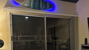 A modern living room with a glass sliding door leading to a balcony, a dark glass coffee table, and a sofa with a patterned throw pillow. - Maui Beach Front Condo Walking Distance to Beaches and Restaurants, ocean views (Kihei)