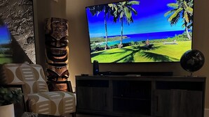 A living room with a flat-screen TV mounted on the wall, a dark wooden entertainment center, a patterned sofa, and a coffee table. - Maui Beach Front Condo Walking Distance to Beaches and Restaurants, ocean views (Kihei)
