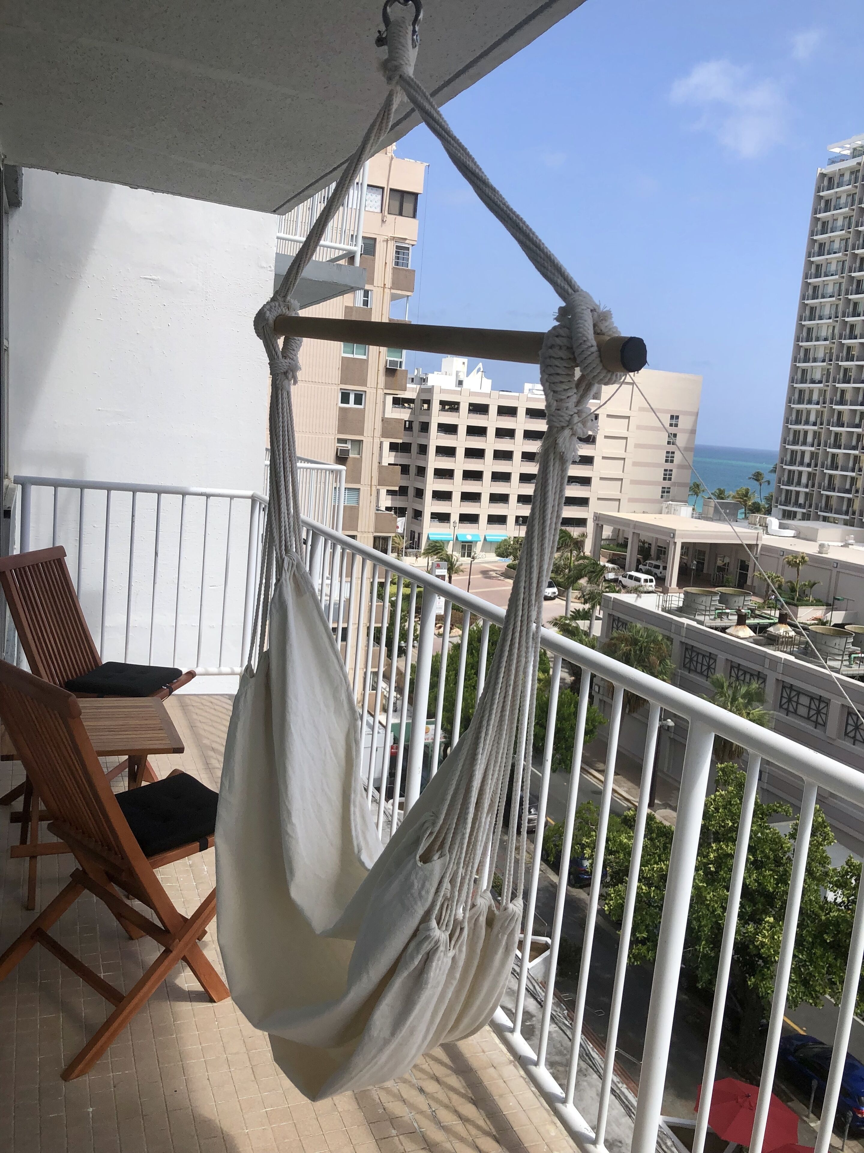 A balcony with a hammock, wooden chairs, and a view of the ocean and buildings.