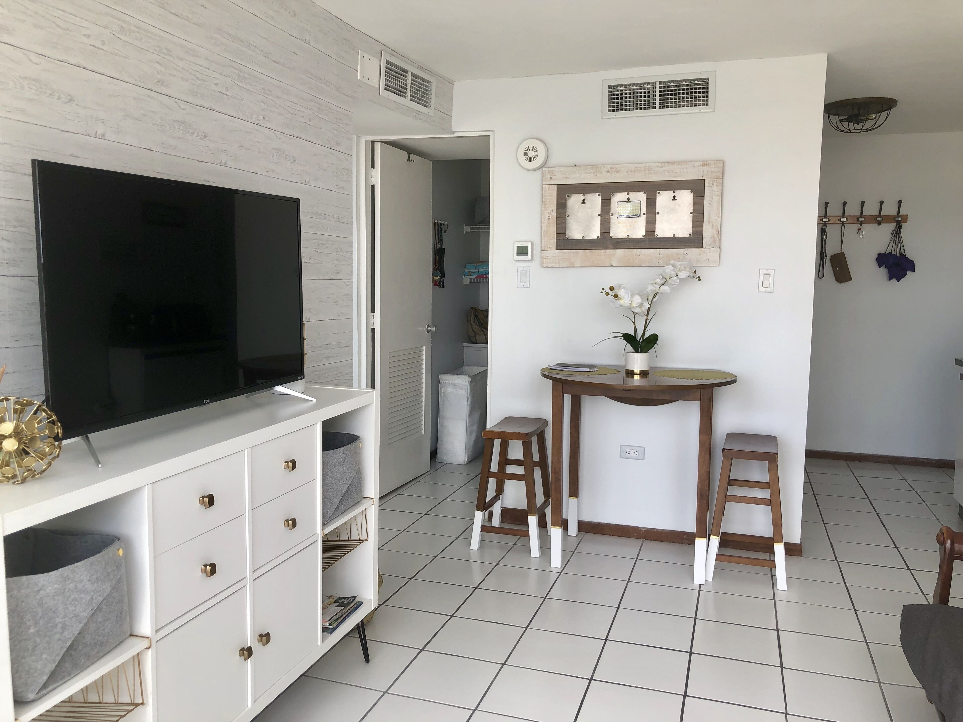 A modern living room with a flat-screen TV, a white entertainment unit, a small round table with two stools, and a decorative wall-mounted shelf.