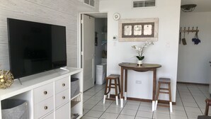 Een moderne woonkamer met een flatscreen televisie, een witte tv-kast, een klein rond tafeltje met twee krukken, en een decoratieve wandplank.