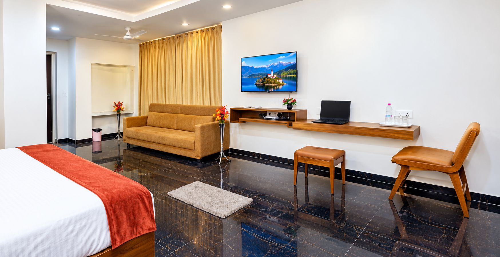 A hotel room with a bed, a sofa, a desk with a laptop, a chair, and a TV displaying a scenic landscape.