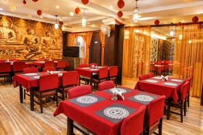 A restaurant with red tablecloths and wooden chairs, a Buddha mural on the wall, and decorative hanging lanterns. - Amaravathi Hotel (Guntur)