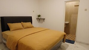 A bedroom with a bed, a nightstand, a small shelf with a plant, and a bathroom door. - Moshi-Moshi - Hostel (Probolinggo)