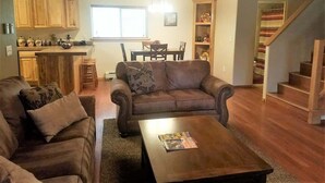 A living room with a brown leather sofa, a wooden coffee table, and a kitchen area with wooden cabinets and a dining table. - Pine Mountain Retreat (Ruidoso)
