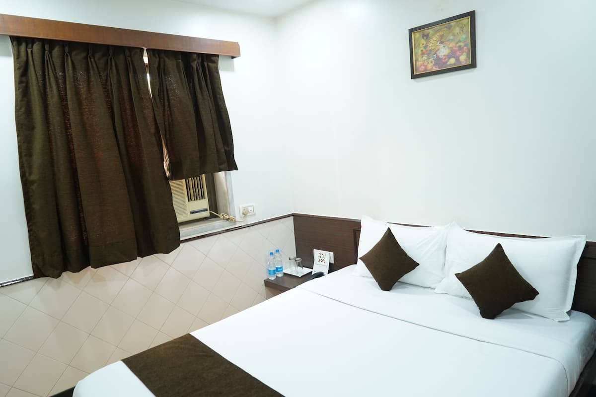 A hotel room with a bed, a nightstand, a window with curtains, a painting on the wall, and a small table with a water bottle and a tissue box.