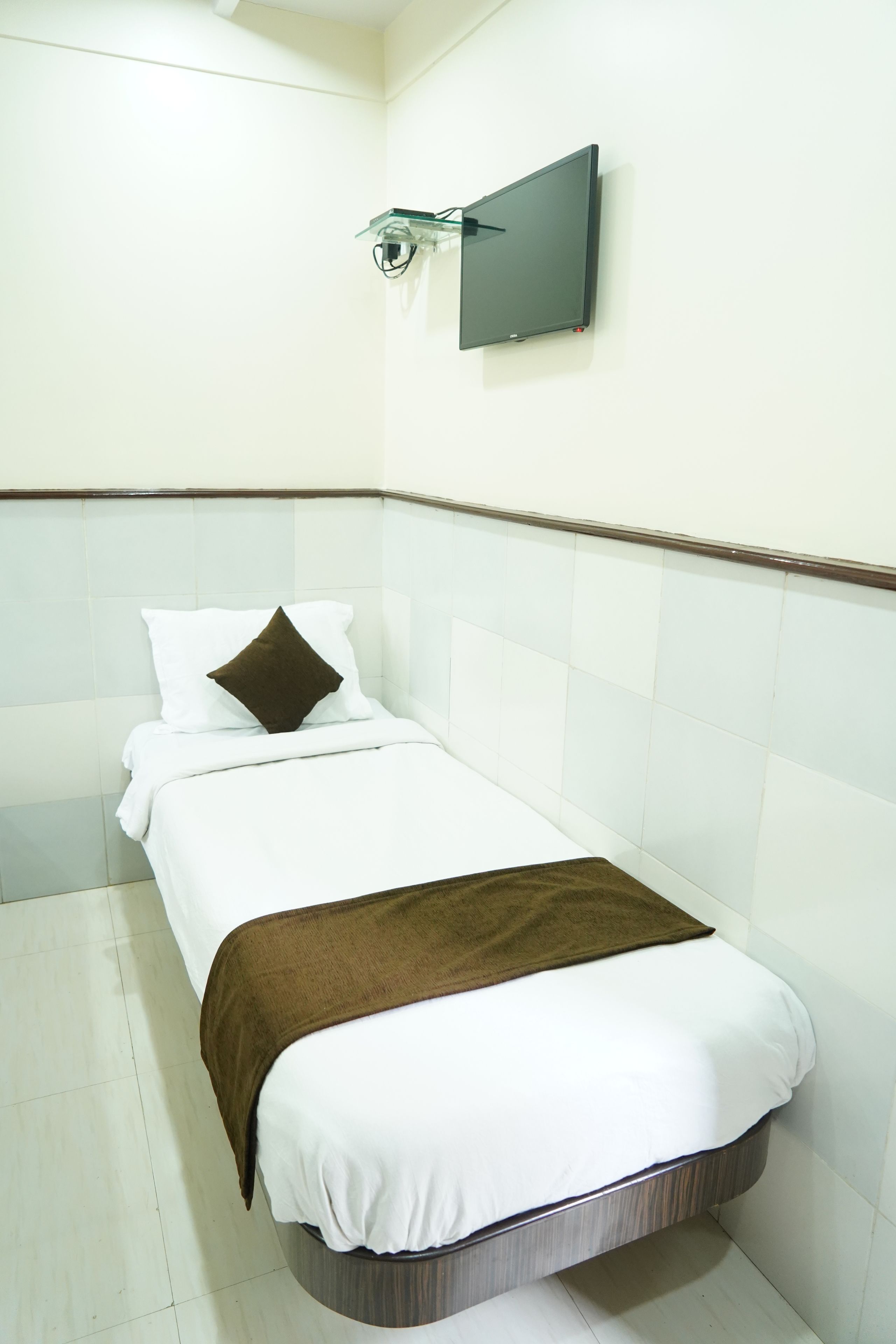 A single bed with white bedding and a brown throw blanket, a flat-screen TV mounted on the wall, and white tiled walls.