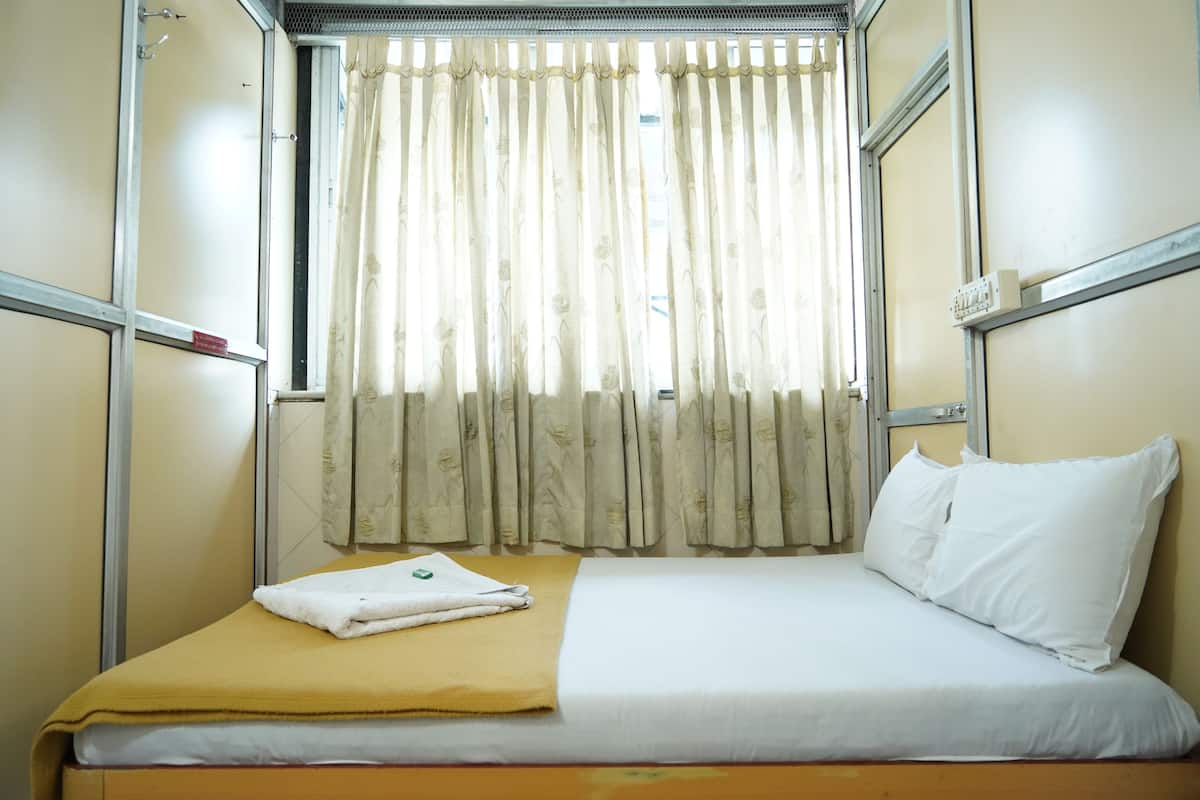 A single bed with a yellow blanket and white pillows in a room with beige walls and a window with curtains.