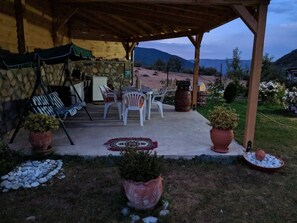 Un patio al aire libre con un muro de piedra, una zona de estar cubierta con sillas y una mesa, plantas en macetas y vistas a montañas al fondo.