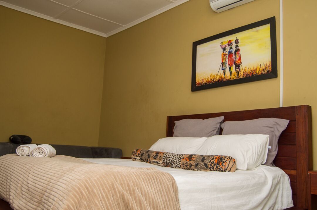 A hotel room with a bed, pillows, a blanket, a painting on the wall, and a small table with a pillow and a rolled towel.