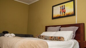 A hotel room with a bed, pillows, a blanket, a painting on the wall, and a small table with a pillow and a rolled towel.