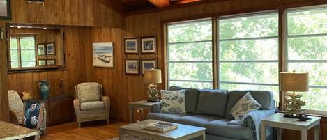 A cozy living room with wooden walls and ceiling, a grey sofa, a coffee table, and a large window with a view of trees.
