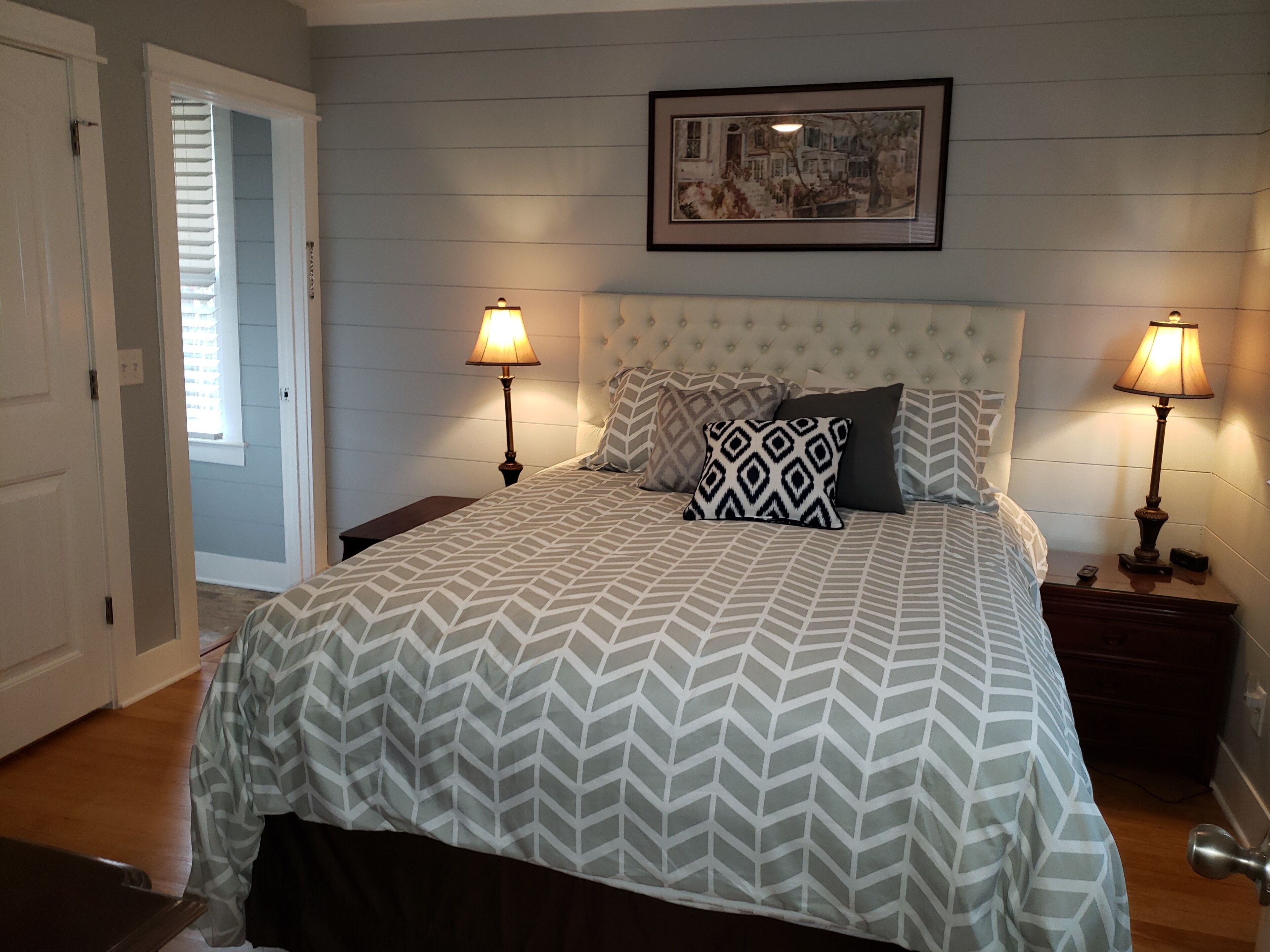 A bedroom with a large bed, two bedside lamps, a framed picture on the wall, and a door leading to another room.