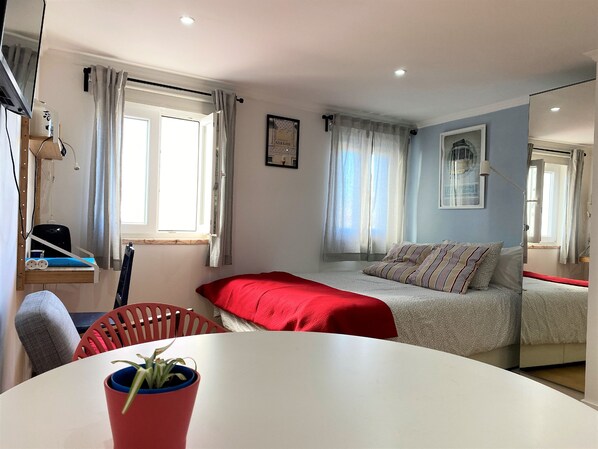 A modern bedroom with a bed, a desk, and a chair. There is a window with curtains, a plant on the desk, and a framed picture on the wall.