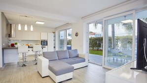 A modern kitchen with white cabinets, a dining area with bar stools, a grey sectional sofa, and a large sliding glass door leading to an outdoor patio. - New elegant Villa EMANUELE in Mondello Beach (Palermo)