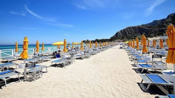 A beach with rows of sun loungers and yellow umbrellas, a clear sky, and a rocky hillside in the background. - New elegant Villa EMANUELE in Mondello Beach (Palermo)