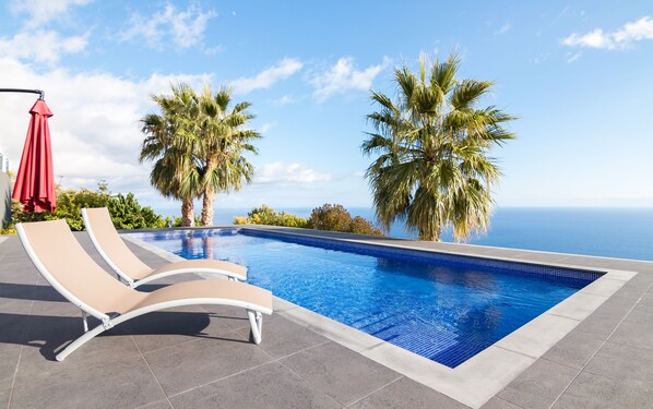 A swimming pool with lounge chairs and palm trees, overlooking a body of water.