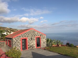 A stone building with a red roof and two red-framed doors, situated on a grassy slope with a view of the sea.
