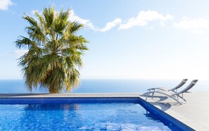 A swimming pool with clear blue water, a palm tree, and lounge chairs on a terrace with a view of the ocean.