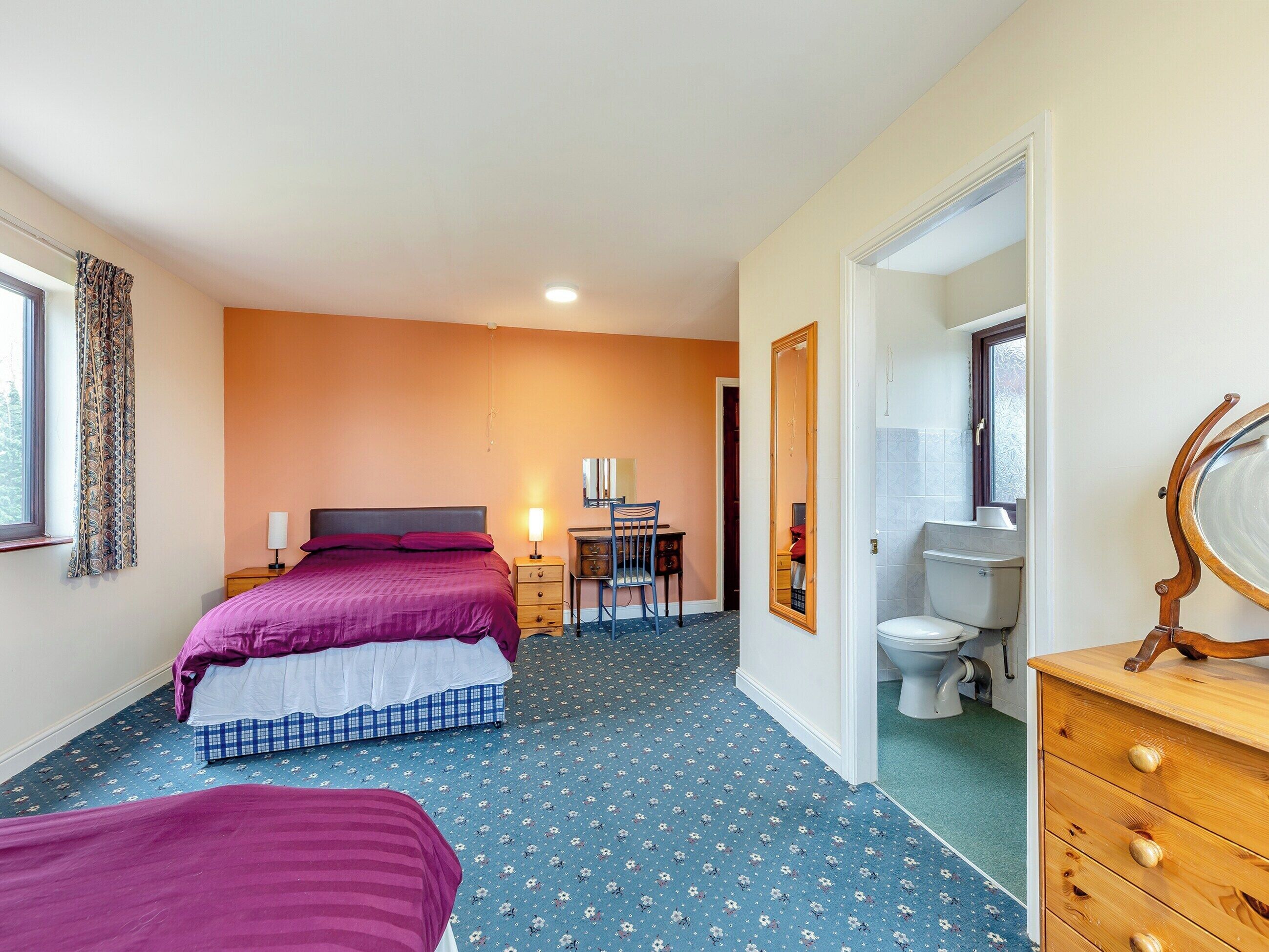 A bedroom with two beds, a desk, a chair, a dresser, and a bathroom with a toilet and mirror.